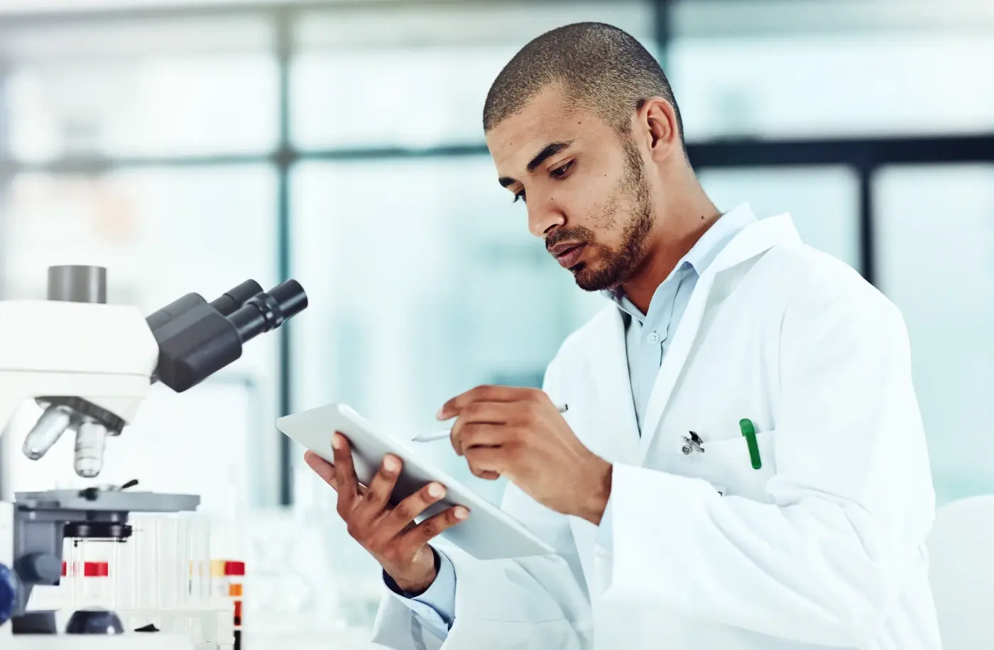 A researcher in a lab coat recording results on a tablet beside a microscope